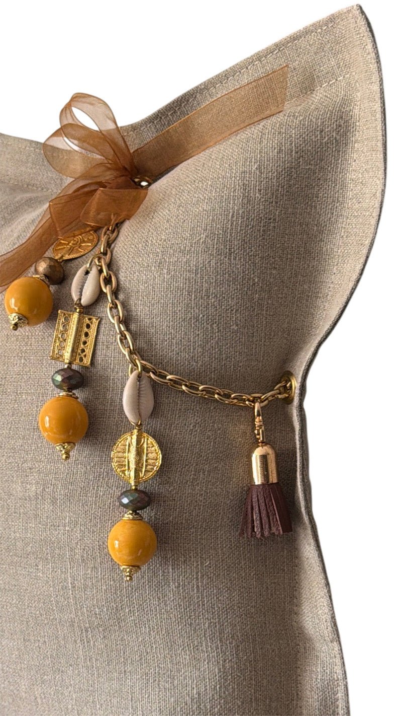 Pampille signature MIYA BALI No.98 avec perles africaines jaunes, cauris et poids Ashanti sur coussin en lin grège.