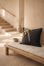Coussin en lin MIYA couleur anthracite avec pampille signature ornée de poids Ashanti et perles, posé sur un banc en bois