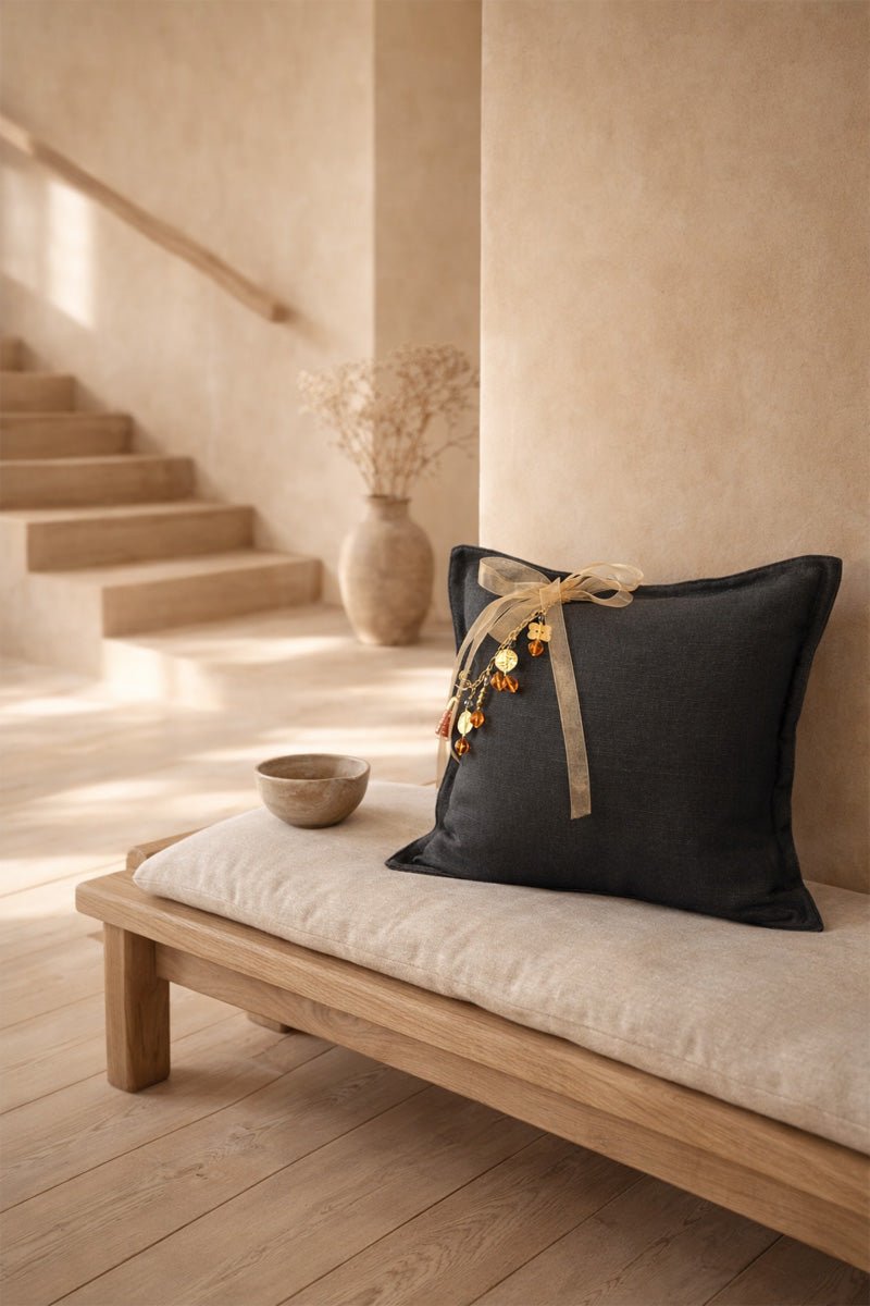 Coussin en lin MIYA couleur anthracite avec pampille signature ornée de poids Ashanti et perles, posé sur un banc en bois