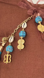 Détail de la pampille signature MIYA MARRAKECH No.49 sur coussin en lin terracotta, associant chaîne dorée, cauris naturels, perles turquoise à motifs floraux, poids Ashanti dorés